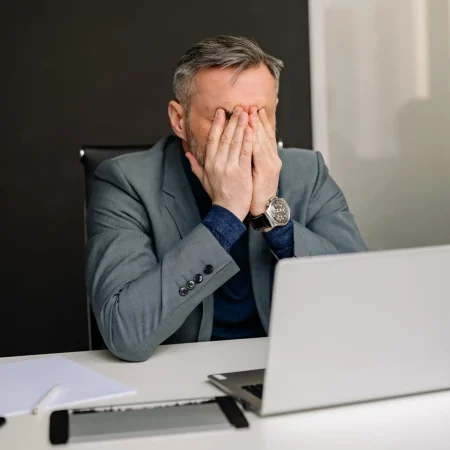 Uomo in abito formale che si copre il volto al computer, simbolo di stanchezza profonda e necessità di fermarsi, supporto